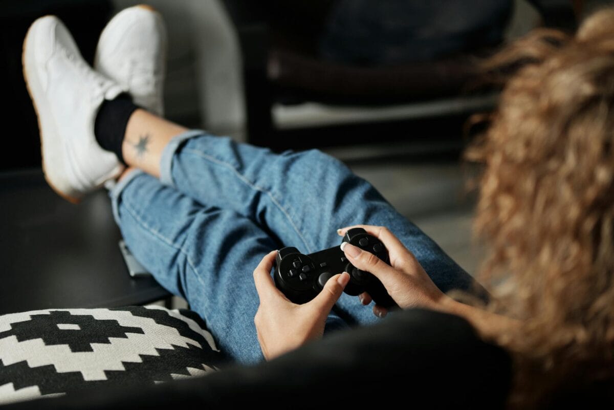 Person sitting on a couch, wearing jeans and white sneakers, holding a game controller with legs crossed on a black and white patterned cushion.