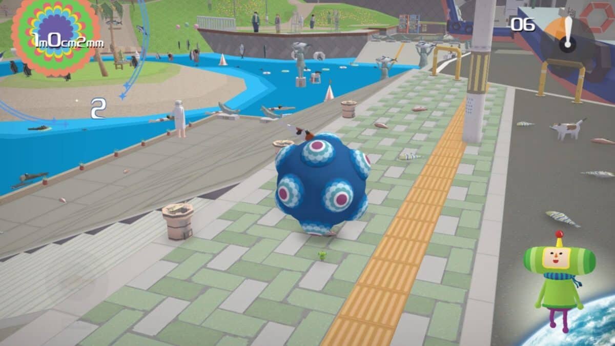 A blue spiked ball rolls on a sidewalk near a river in a colorful video game world, with small objects and characters scattered around.