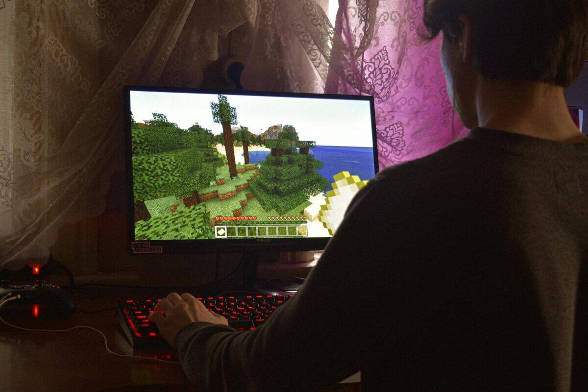 A person sits at a desk playing Minecraft on a computer, using a keyboard and mouse, with lace curtains and pink lighting in the background.