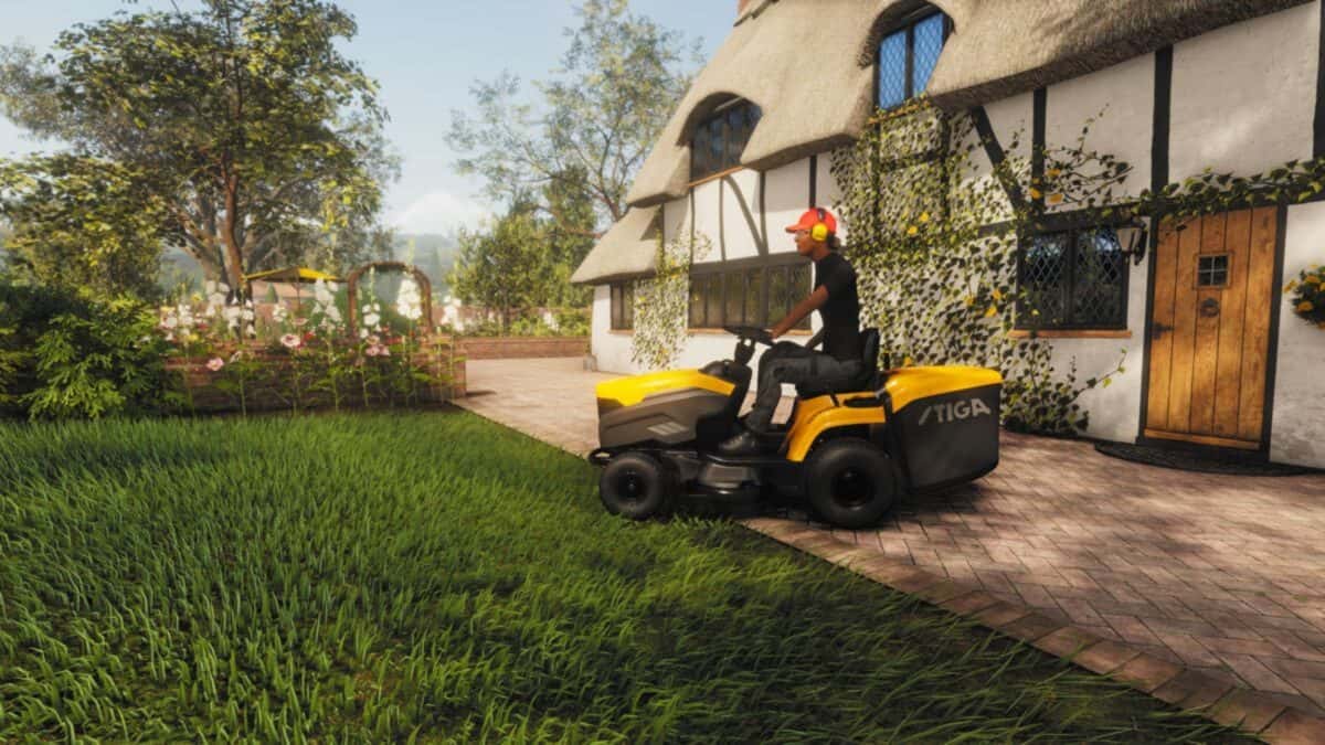 A person wearing ear protection operates a yellow ride-on lawn mower on grass near a house with a thatched roof and timber framing.