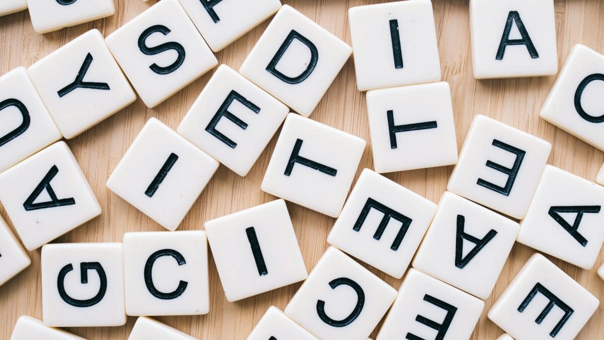 Scattered white letter tiles with black uppercase letters on a light wooden surface, arranged in no particular order.