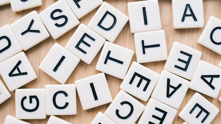 Scattered white letter tiles with black uppercase letters on a light wooden surface, arranged in no particular order.