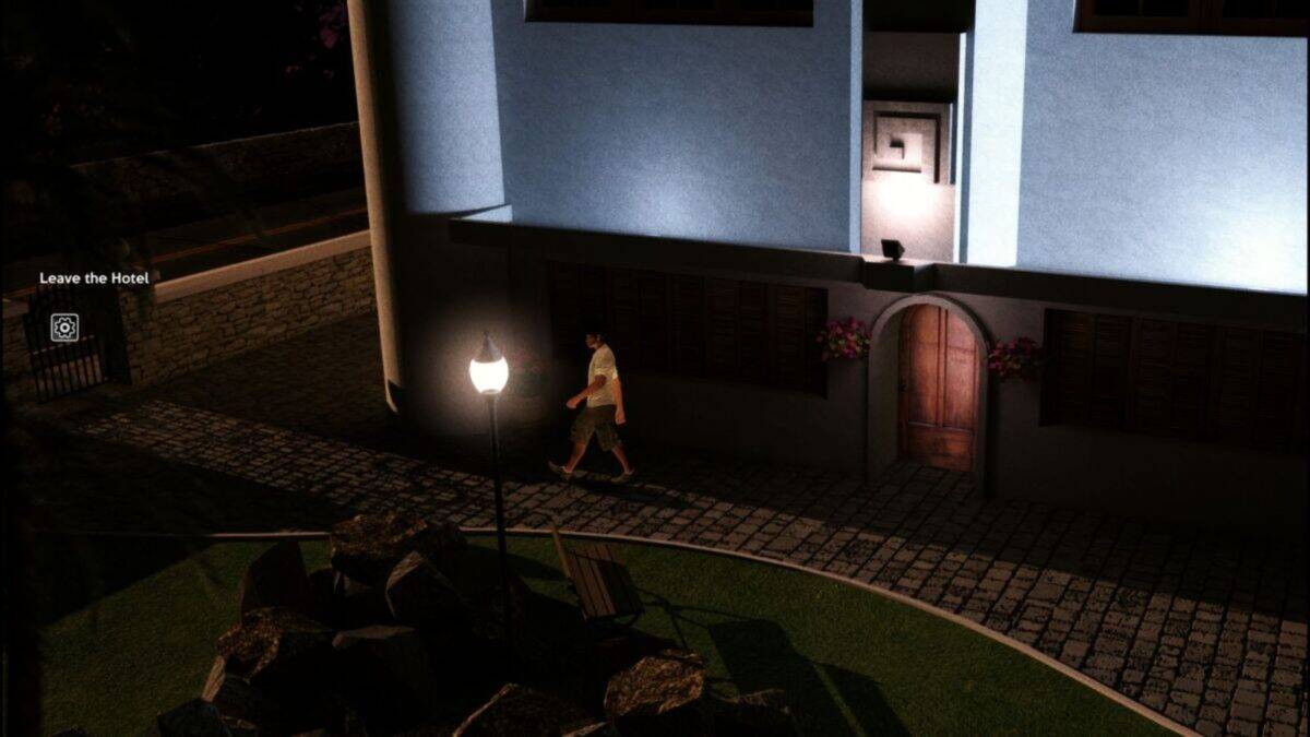 A man walks on a cobblestone path beside a blue building at night, illuminated by a streetlamp and surrounded by a garden area.