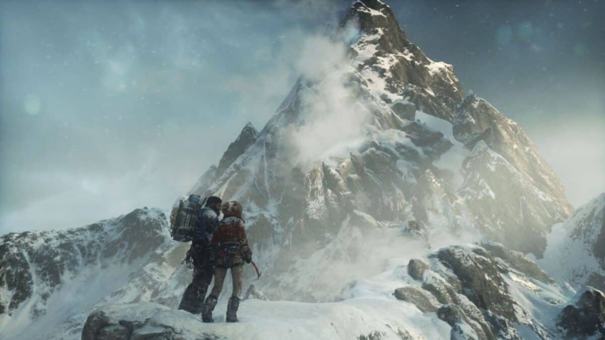 Two climbers wearing winter gear stand on a snowy ridge, looking up at a tall, rugged, snow-covered mountain peak under a cloudy sky.